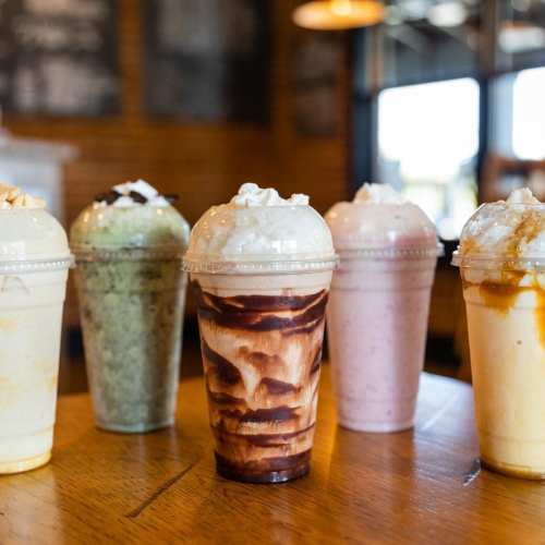 Five colorful milkshake cups with whipped cream, arranged on a wooden table in a cafe setting.