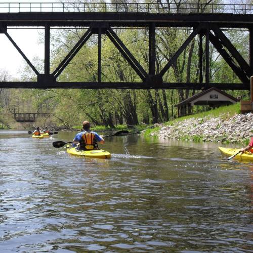 Two kayakers paddle on a calm river beneath a bridge, surrounded by lush greenery and a peaceful landscape.