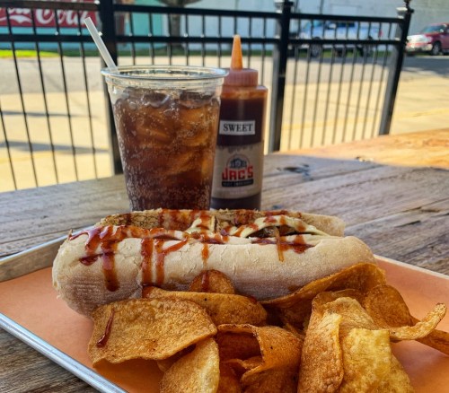 A hot dog topped with sauce, served with potato chips and a drink on a wooden table.