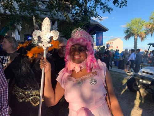 A woman in a pink wig and tiara holds a decorative staff, smiling at a festive outdoor event with palm trees in the background.