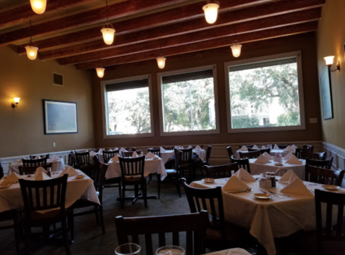 Cozy restaurant interior with wooden beams, tables set with white linens, and large windows overlooking greenery.