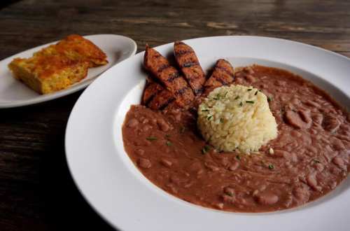 A plate of rice and beans with grilled sausage, garnished with herbs, alongside a slice of cornbread.