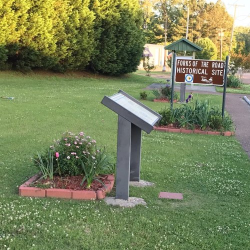 Sign and informational display at the Forks of the Road Historical Site, surrounded by grass and flowers.