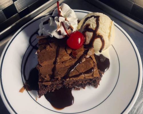 A slice of chocolate brownie topped with whipped cream, a cherry, and a scoop of vanilla ice cream drizzled with chocolate sauce.