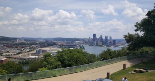 A panoramic view of a city skyline with a river, surrounded by greenery and a partly cloudy sky.