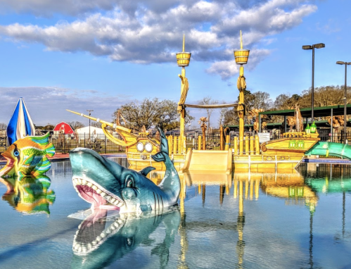 Colorful water park scene featuring a large shark float and pirate ship structures in a sunny outdoor setting.