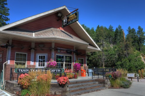A charming train station with a pink exterior, surrounded by colorful flowers and greenery under a clear blue sky.