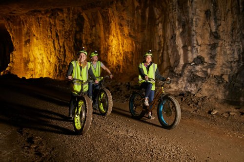 Three people in reflective vests and helmets ride fat bikes through a dimly lit cave with rocky walls.