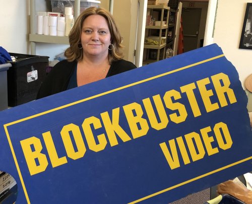 A woman holds a large Blockbuster Video sign, smiling in a store setting.