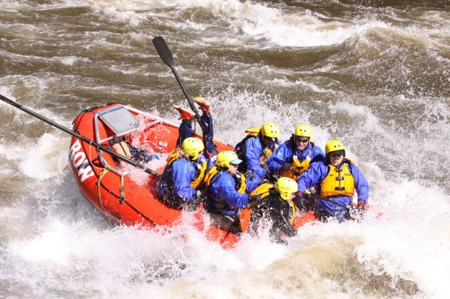 A group of eight people in a red raft navigating turbulent whitewater rapids, all wearing helmets and life jackets.