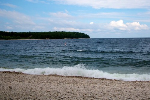 A serene beach scene with gentle waves, a rocky shore, and a paddleboarder in the distance under a partly cloudy sky.