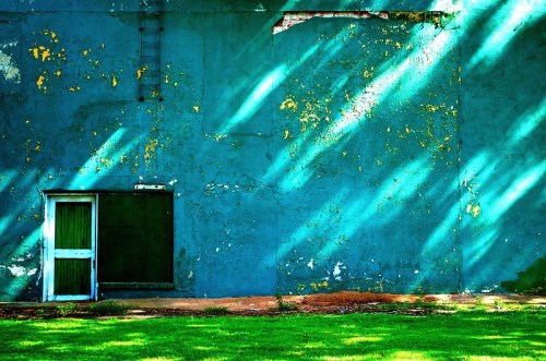 A weathered blue wall with peeling paint, featuring a green door and window, illuminated by diagonal sunlight.