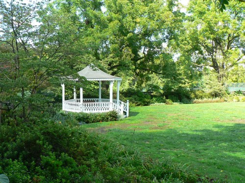 A white gazebo surrounded by lush greenery and trees in a sunny park setting.