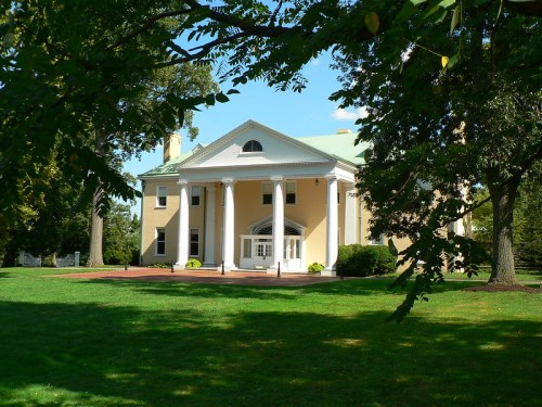 A large yellow house with white columns, set in a green landscape under a clear blue sky.