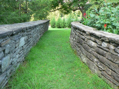 A narrow path between two stone walls, surrounded by lush green grass and trees.