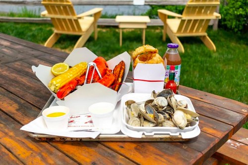 A seafood feast featuring lobster, corn, clams, and a drink on a wooden table in a grassy outdoor setting.
