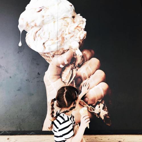 A child with pigtails holds an ice cream cone, standing next to a large mural of a hand holding an ice cream cone.