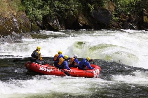 A group of six people in a red raft navigating through whitewater rapids surrounded by lush greenery.