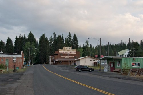 A quiet rural street with a few buildings, surrounded by tall trees under a cloudy sky. A vintage car is parked nearby.