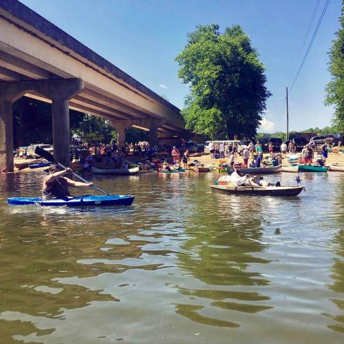 A sunny day at a river with people kayaking and relaxing under a bridge, surrounded by trees and a lively crowd.