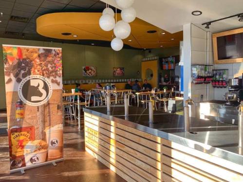 A bright, modern café interior with a beverage station, seating area, and a colorful promotional banner.