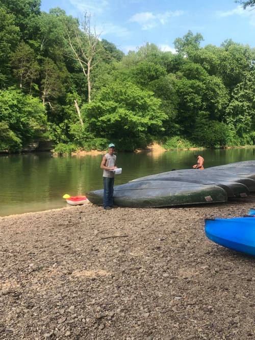 A person stands on a riverbank near kayaks, surrounded by lush green trees and a calm water scene.