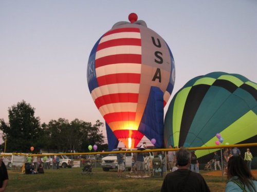 A colorful hot air balloon with a USA design is illuminated at dusk, surrounded by spectators and other balloons.