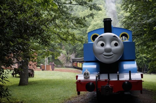 A cheerful blue train with a smiling face stands on a green track surrounded by trees and a scenic landscape.