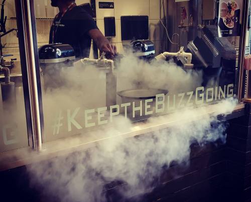 A person stands behind a counter with fog and a "#KeepTheBuzzGoing" sign, suggesting a lively atmosphere.
