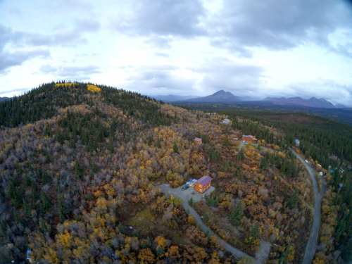 Aerial view of a forested landscape with a small orange building surrounded by autumn foliage and distant mountains.