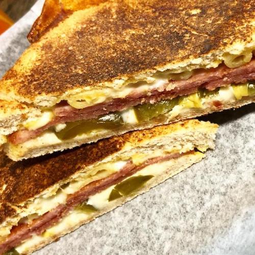 Close-up of a toasted sandwich with layers of ham, cheese, pickles, and macaroni, cut in half on parchment paper.