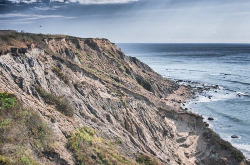 A rugged coastline with steep cliffs, waves crashing below, and a clear sky in the background.