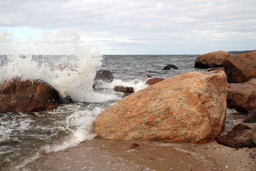 Waves crashing against large rocks on a sandy beach under a cloudy sky.
