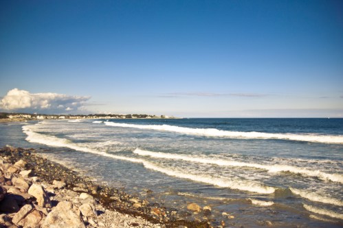 A serene beach scene with gentle waves, rocky shore, and a clear blue sky in the background.