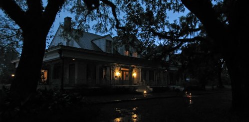 A dimly lit historic house surrounded by trees at dusk, with warm lights glowing from the windows.