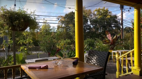A cozy outdoor dining area with a wooden table, chairs, and lush greenery in the background under a blue sky.