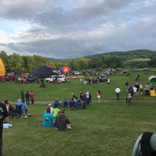 A lively outdoor event with colorful hot air balloons, people gathered on grass, and mountains in the background.
