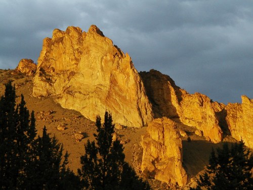 Golden rock formations illuminated by sunlight against a dramatic cloudy sky. Dark trees in the foreground.