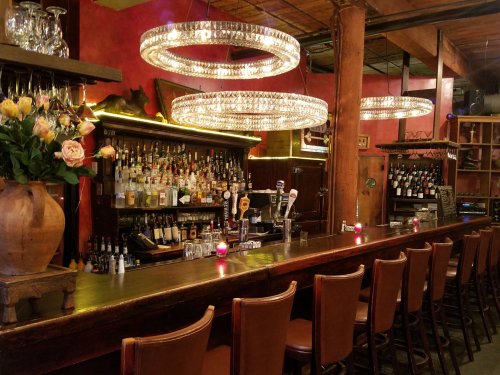 A cozy bar with a wooden counter, elegant chandeliers, and shelves stocked with various bottles and wine.