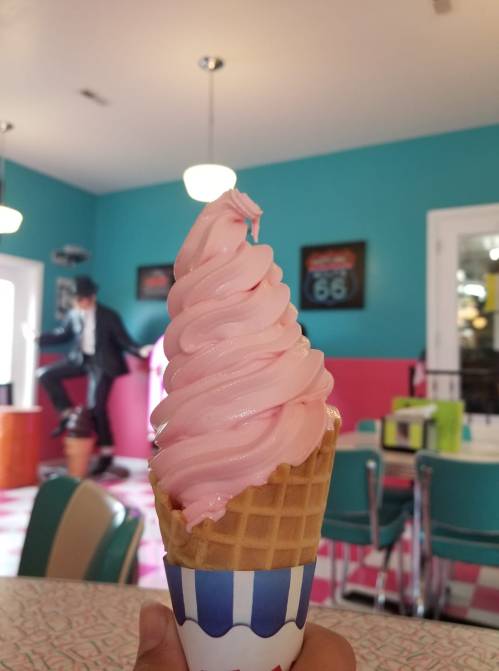A hand holds a cone of pink soft serve ice cream in a retro diner with colorful decor.