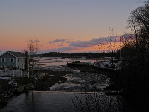 A serene river scene at sunset, with a house on the left and colorful clouds reflecting on the water.