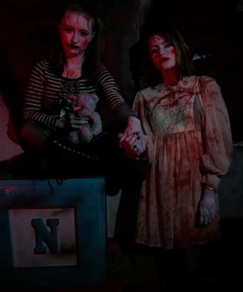 Two girls in eerie costumes, one in stripes holding a stuffed toy, both with painted faces and bloodstains, in a dimly lit setting.