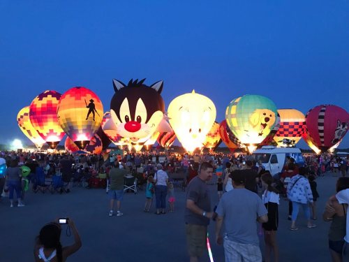 Colorful hot air balloons, including cartoon characters, illuminate the night sky as a crowd enjoys the festival.