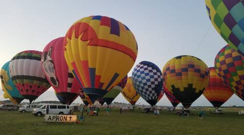 Colorful hot air balloons fill the sky at a festival, with spectators and vehicles on a grassy field below.