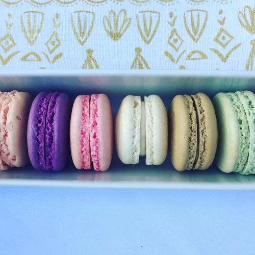 A row of colorful macarons in a white box, featuring purple, pink, white, beige, and green varieties.
