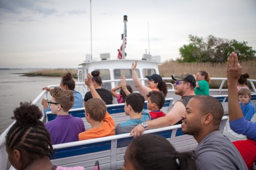 A diverse group of children and adults on a boat, raising their hands while enjoying a scenic ride on the water.
