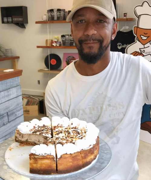A man in a cap smiles while holding a cheesecake topped with whipped cream and nuts in a cozy shop setting.