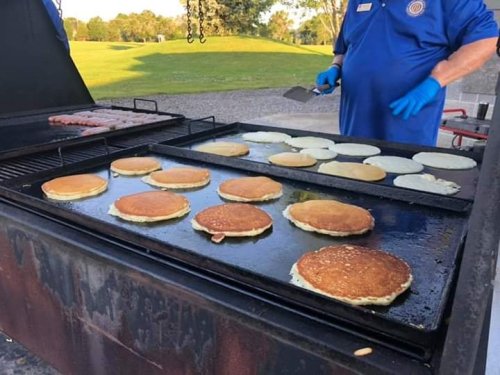 A grill with pancakes cooking on one side and sausages on the other, with a person in blue gloves flipping pancakes.