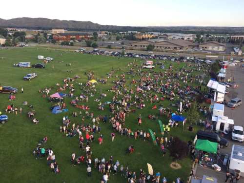 Aerial view of a large crowd gathered on a grassy field for an outdoor event, with tents and vehicles in the background.