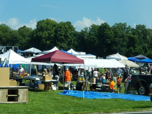 A bustling outdoor market with various tents, vendors, and people browsing among tables of goods on a sunny day.